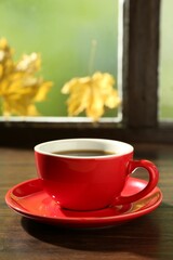 Red cup of aromatic coffee and saucer on wooden sill near window