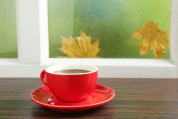 Red cup of aromatic coffee and saucer on wooden sill near window, closeup
