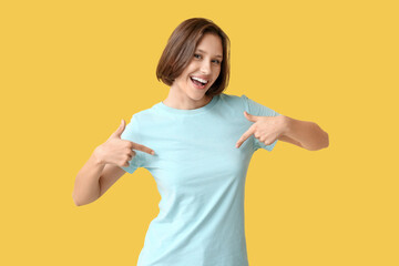 Young woman pointing at her blank t-shirt on yellow background