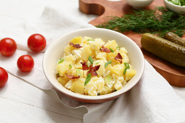 Bowl of tasty potato salad with bacon, green onion, tomatoes and pickled cucumbers on white wooden background