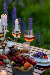 Two glasses of rose wine, flowers in a vase, silverware, fruits. Sunset, golden hour, atmospheric candlelight. Romantic table decor for a loving couple on blooming meadow with purple lupines