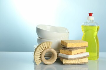 Stack of sponges, brush, bottle with detergent and dishware on light table, closeup. Space for text