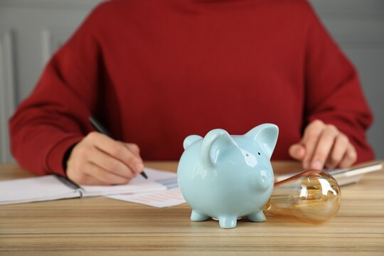 Man taking notes while using calculator at wooden table, focus on piggy bank and light bulb. Energy saving concept