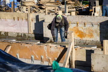 Construction worker in toolbelt and gloves placing metal stakes in freshly poured cement foundation footings, home building job site on a sunny winter day
