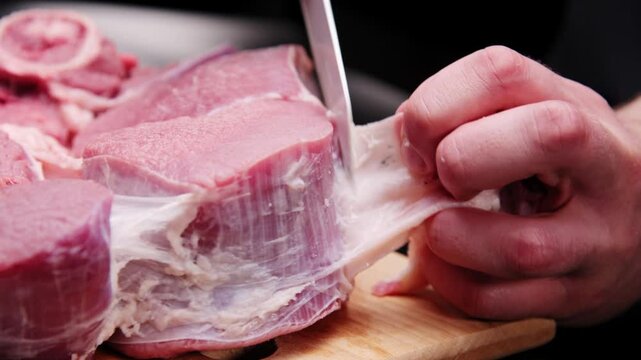 A butcher removes the fascia from a beef steak with a large knife in a dark room