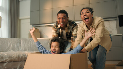 Happy African American family celebrate relocation new home joyful mother father run push cardboard box with little child boy kid ride inside cheerful parents son playing fun moving day loan mortgage