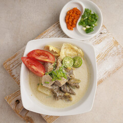 a bowl of Indonesian cuisine or Soto Betawi, a creamy soup served with sliced meat, vegetables and crackers, in white bowl