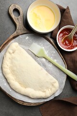 Uncooked calzone pizza, sauce and egg yolk on grey table, flat lay