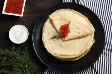 Tasty crepes with red caviar, dill and sauce on wooden table, flat lay
