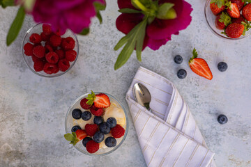Top view of a summer dessert with vanilla ice cream, fresh strawberries, blueberries, and raspberries in a glass bowl on a textured surface next to a white napkin, spoon, and vibrant pink peonies