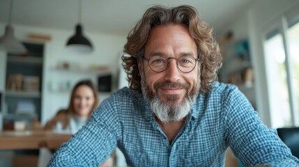 A lively man with round glasses and curly hair beams with a joyful smile, wearing a checkered shirt, seated in a bright and neatly arranged indoor space.