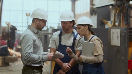 Industrial engineer wearing glasses, formal shirt and hard hat reviewing documents and discussing work plan with male and female factory workers