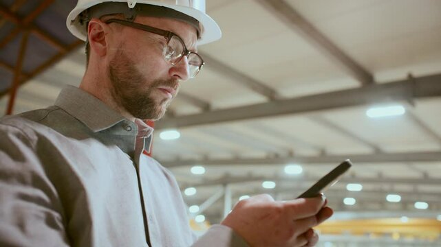 Focused engineer in hard hat, formalwear and glasses standing in industrial plant and using mobile phone during workday. Low angle view