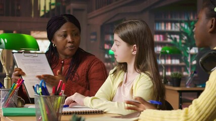 African american professor mentoring diverse schoolgirls in the library, learning together in a library filled with curriculum based materials. Woman tutoring pupils on literature. Camera A.
