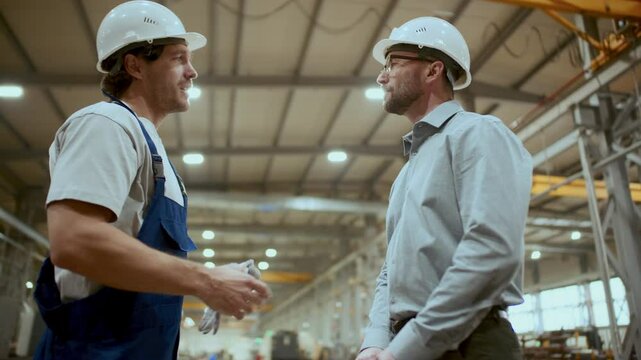 Engineer in hardhat shaking hands with industrial worker, discussing manufacturing processes and equipment with him during meeting in factory. Low angle view