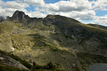A fragment of a wide lake lying in a low mountain valley surrounded by pointed rocks under a cloudy summer sky.