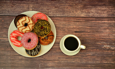Large plate with assortment of fresh donuts glazed with colorful decor on wooden table, cup of hot black coffee on saucer nearby.