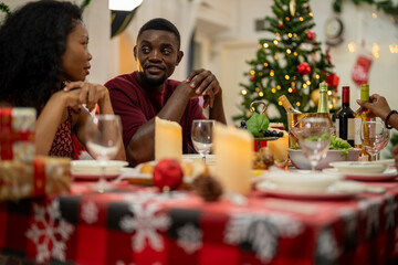 A joyful family gathers around the dinner table celebrating Christmas. The father, dressed in a Santa hat, gives a gift to the child, while everyone smiles with warmth and festive decorations.
