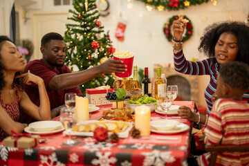 A joyful family gathers around the dinner table celebrating Christmas. The father, dressed in a Santa hat, gives a gift to the child, while everyone smiles with warmth and festive decorations.