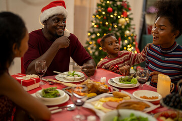 A joyful Christmas celebration with friends gathered around a festive dining table. Featuring a man in a Santa hat, a decorated Christmas tree, delicious food, and warm holiday cheer.