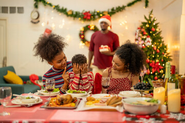 A joyful Christmas celebration with friends gathered around a festive dining table. Featuring a man in a Santa hat, a decorated Christmas tree, delicious food, and warm holiday cheer.