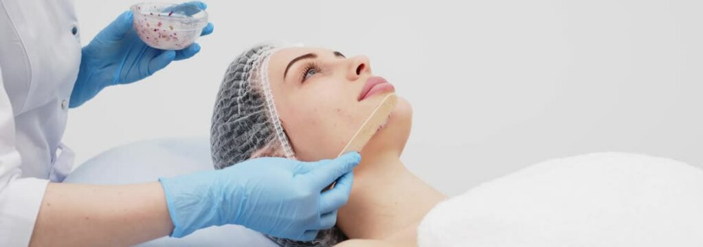 Close-up of a cosmetologist&rsquo;s hands applying an alginate mask with rose petals to a young woman&rsquo;s face in a professional beauty salon. Shot on anamorphic lens in 2.39