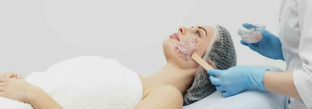 Close-up of a cosmetologist&rsquo;s hands applying an alginate mask with rose petals to a young woman&rsquo;s face in a professional beauty salon. Shot on anamorphic lens in 2.39
