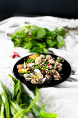 Garlic clams and mussels are presented on a black plate, surrounded by green herbs and chili.