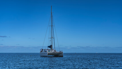 Fototapeta premium A white yacht in the blue ocean. Silhouettes of people on the deck of a catamaran. A tall mast and rigging against a clear azure sky. Copy Space. Mauritius.