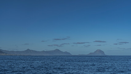 The coast of a tropical island is visible from the ocean. Picturesque silhouettes of mountains against the background of blue sky and clouds. Ripples on the surface of the water. Mauritius. Copy Space