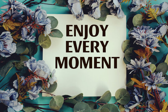 Enjoy Every Moment text with flowers frame decoration top view on blue background, Inspirational and motivational quote