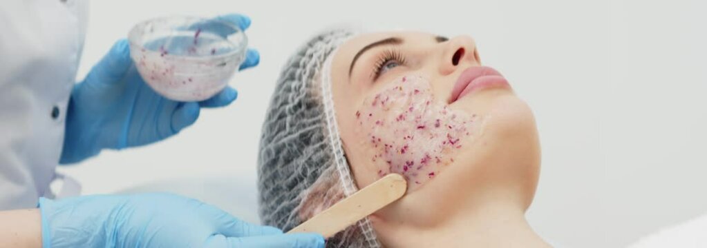 Close-up of a cosmetologist&rsquo;s hands applying an alginate mask with rose petals to a young woman&rsquo;s face in a professional beauty salon. Shot on anamorphic lens in 2.39