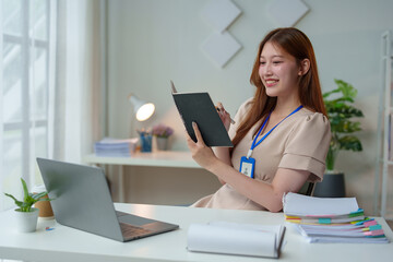 Happy independent young Asian female business owner sitting at work holding a notebook Jot down important information, reflect on ideas, and use your laptop at your online meeting desk.