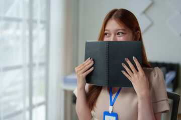 Happy independent young Asian female business owner sitting at work holding a notebook Jot down important information, reflect on ideas, and use your laptop at your online meeting desk.
