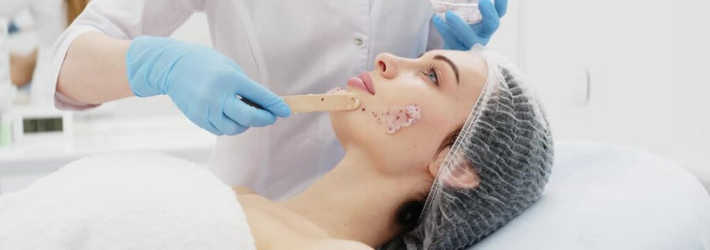 Close-up of a cosmetologist&rsquo;s hands applying an alginate mask with rose petals to a young woman&rsquo;s face in a professional beauty salon. Shot on anamorphic lens in 2.39