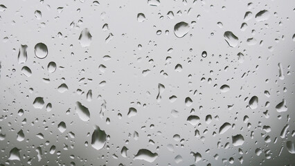 Rain Droplets on Glass - Close-up view of raindrops scattered across a glass surface. The background is softly blurred with a grayish tone, emphasizing the shapes and clarity of the water droplets.