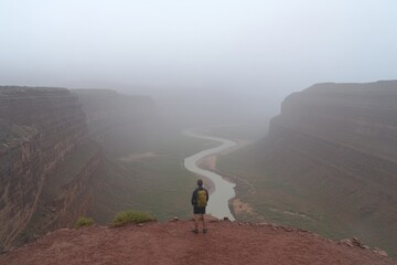 Obraz premium A lone backpacker standing on a cliff edge, staring at a winding river snaking through a vast canyon landscape