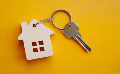 A keyring holds a house-shaped charm and a single key, resting against a bright yellow background. The charm is made of light wood, featuring small square window cutouts