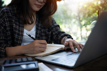 A woman is writing in a notebook while using a laptop. She is focused on her work and she is in a...
