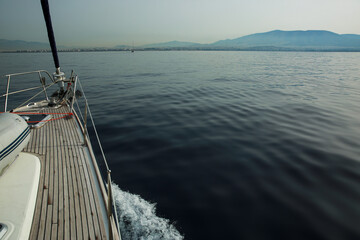 A serene view from the deck of a yacht sailing on calm waters, with distant mountains and the faint skyline of Greece visible under a pale, hazy sky