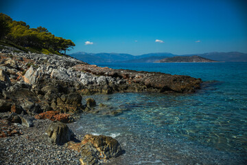 A stunning rocky coastline in Greece with crystal-clear turquoise waters, rugged cliffs, and lush green trees under a bright blue sky
