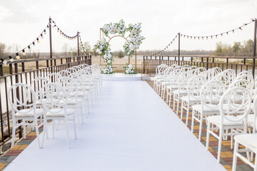 Fototapeta premium Outdoor wedding ceremony. Beautiful and stylish wedding arch decorated with white rose flowers with white chairs standing on the background of water. Day of celebration