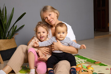 Naklejka premium A grandmother sits on a play mat surrounded by her two grandchildren, smiling and engaging in playful activities. The warm atmosphere highlights family bonding and joy
