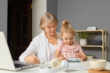 A grandmother and her young granddaughter sit at a table, engaged in a shared activity. The girl interacts with a device while her grandmother observes with joy, creating a warm family moment