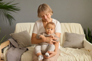 A loving grandmother sits on a soft couch, holding her cute grandson. Morning light fills the cozy living room, creating a warm atmosphere filled with tenderness and connection