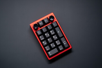 Numpad mechanical keyboard in vibrant red with white on black keycaps, featuring a sleek rotary knob at the top. Top view. Positioned in center on a matte black surface.