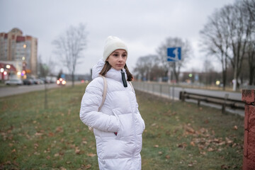 Portrait of a young beautiful girl in a white down jacket in the city in cloudy weather.