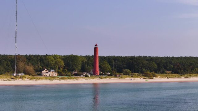 Front aerial view of an orange Lighthouse watchtower at forested beach coastline, natural landscape at daylight, drone shot