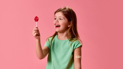 Laughing little girl admiring red lollipop, playful and bright atmosphere, bandage on arm visible against pink studio background. Concept of healthcare, protection, immunity, prevention.