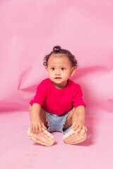 portrait of an 18 month old curly haired girl sitting with focused gaze and stretching her legs isolated on pink